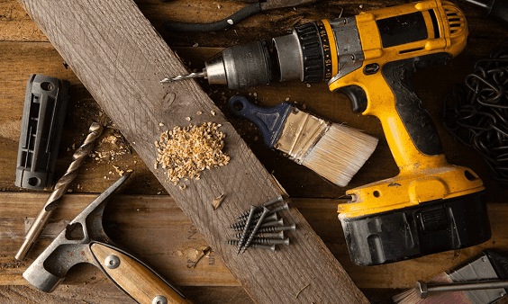 A cordless drill, wooden plank, hammer, nails, and other tools scattered on a dark wooden surface.