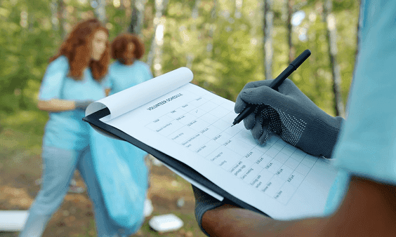 Volunteer writing on a clipboard while others clean up a park in the background.