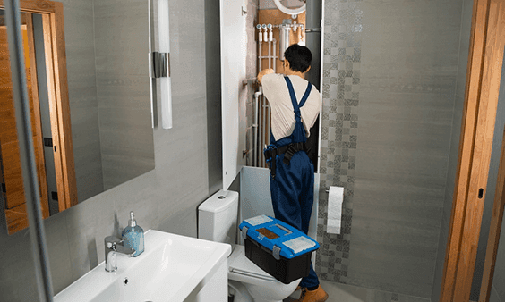 A plumber in blue overalls repairs pipes behind a toilet in a modern bathroom.