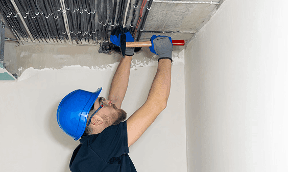 Man in blue hard hat works on ceiling electrical conduits with a tool.