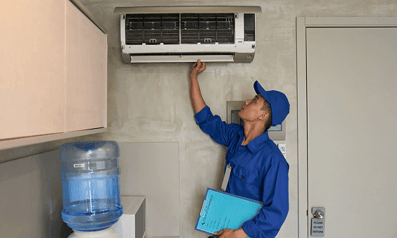 Man in blue uniform inspecting an air conditioner unit mounted on a wall.