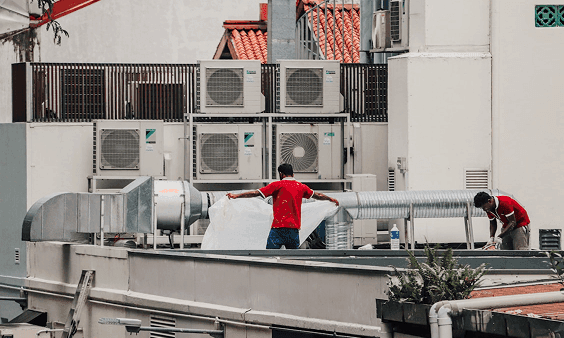 Two workers on a cluttered rooftop with many air conditioning units, one stretching a white sheet.
