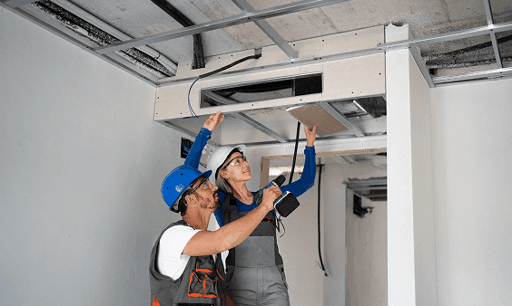 Two construction workers install drywall panels onto a ceiling framework.